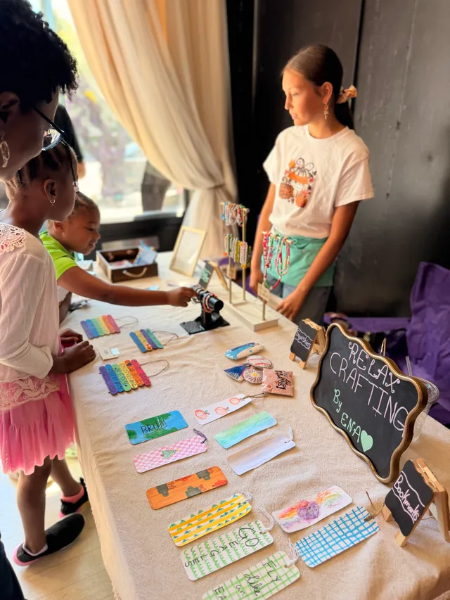 Young vendor stands behind a table of hand-painted bookmarks and bracelets as children and an adult browse.