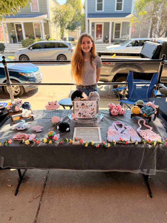 Young vendor smiling behind a table of pink, black and gray crocheted animals, bags and a letter board sign