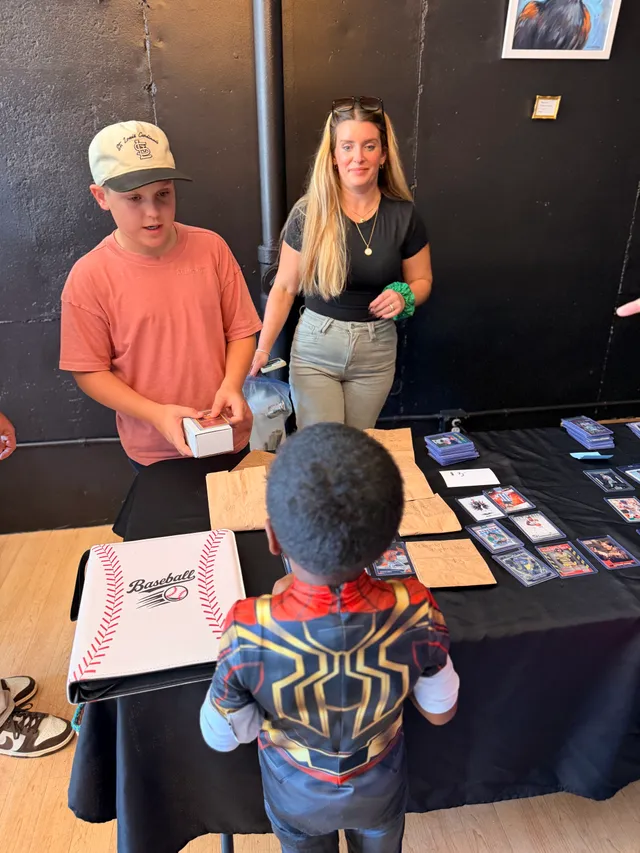 Boy in St. Louis Cardinals cap holds a card box at a trading card table as a child in a Spider-Man costume shops