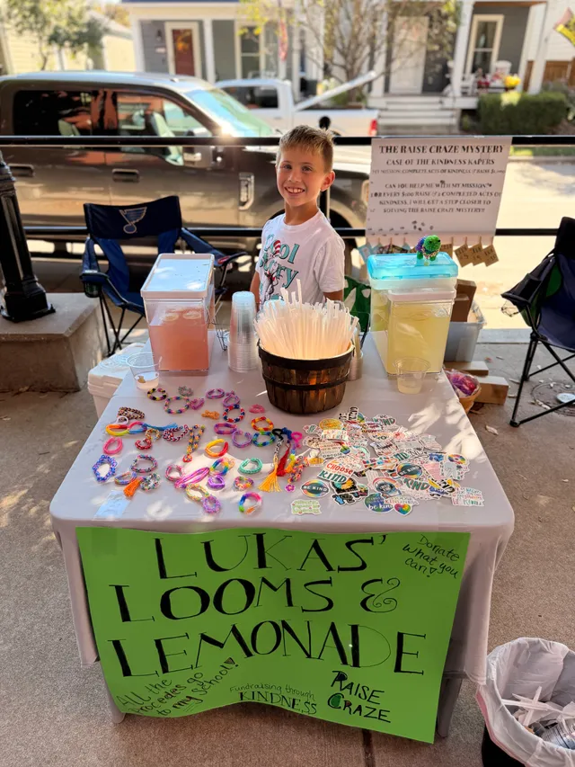Smiling boy behind a table of loom bracelets, stickers and pink and yellow lemonade, with a green Lukas' Looms & Lemonade sign