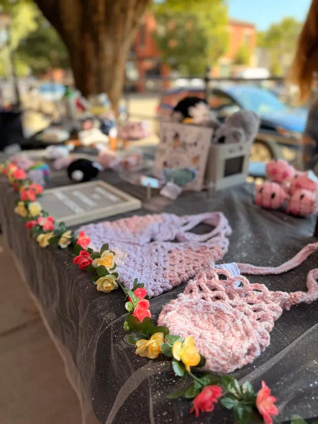Vendor table with pink and lavender crocheted bags, a garland of artificial roses, and crocheted plush toys on dark tulle