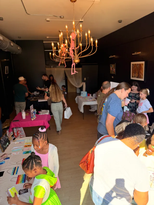 Kids and adults browse vendor tables of crafts in a black-walled room under a gold chandelier with pink skeleton decorations