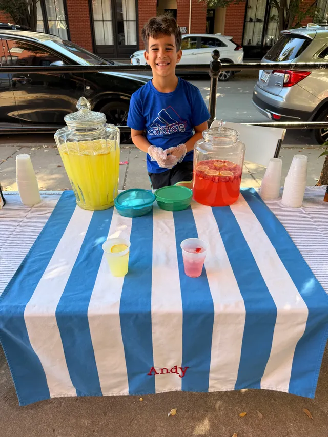 Smiling boy in blue shirt and plastic gloves behind a table with jars of yellow lemonade and pink drink on a blue striped cloth