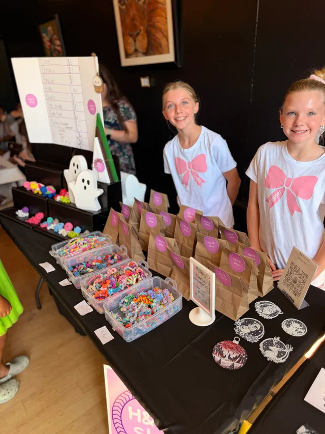 Two girls in white shirts with pink bows smile behind a table of rubber band bracelets, paper goody bags and ornaments.