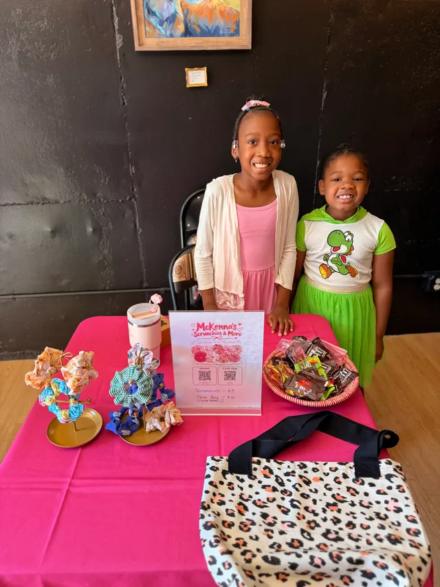 Two smiling girls stand behind a pink-covered table with scrunchie displays, a McKenna's Scrunchies & More sign, candy and a tote bag.