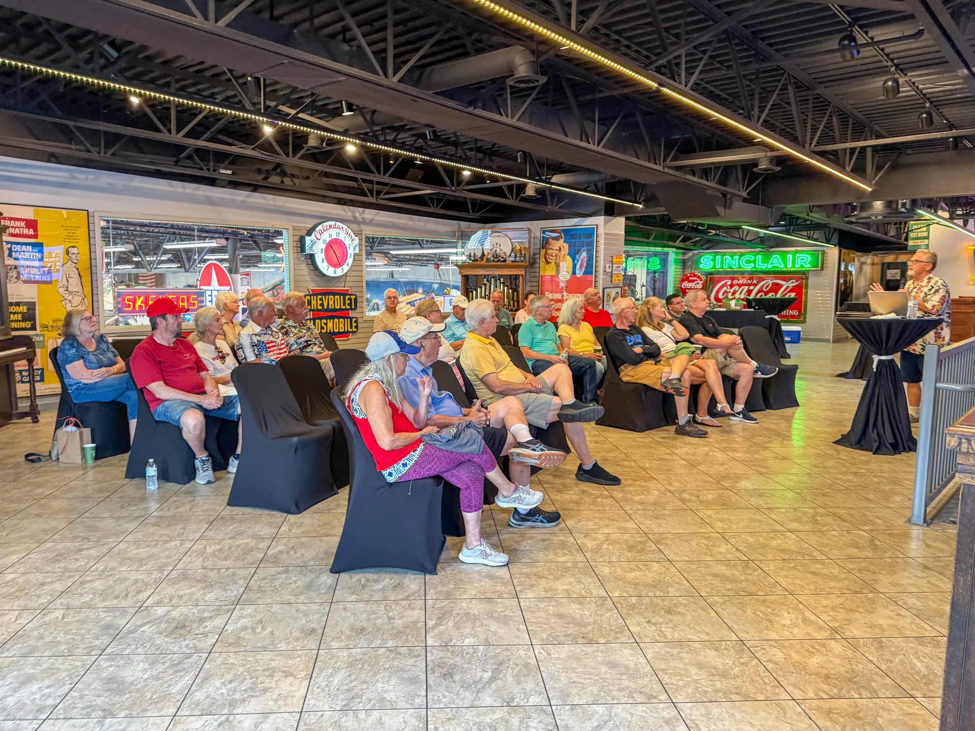 Community members seated among vintage signs at Fast Lane Classic Cars listening to historian Tom Eyssell's lecture