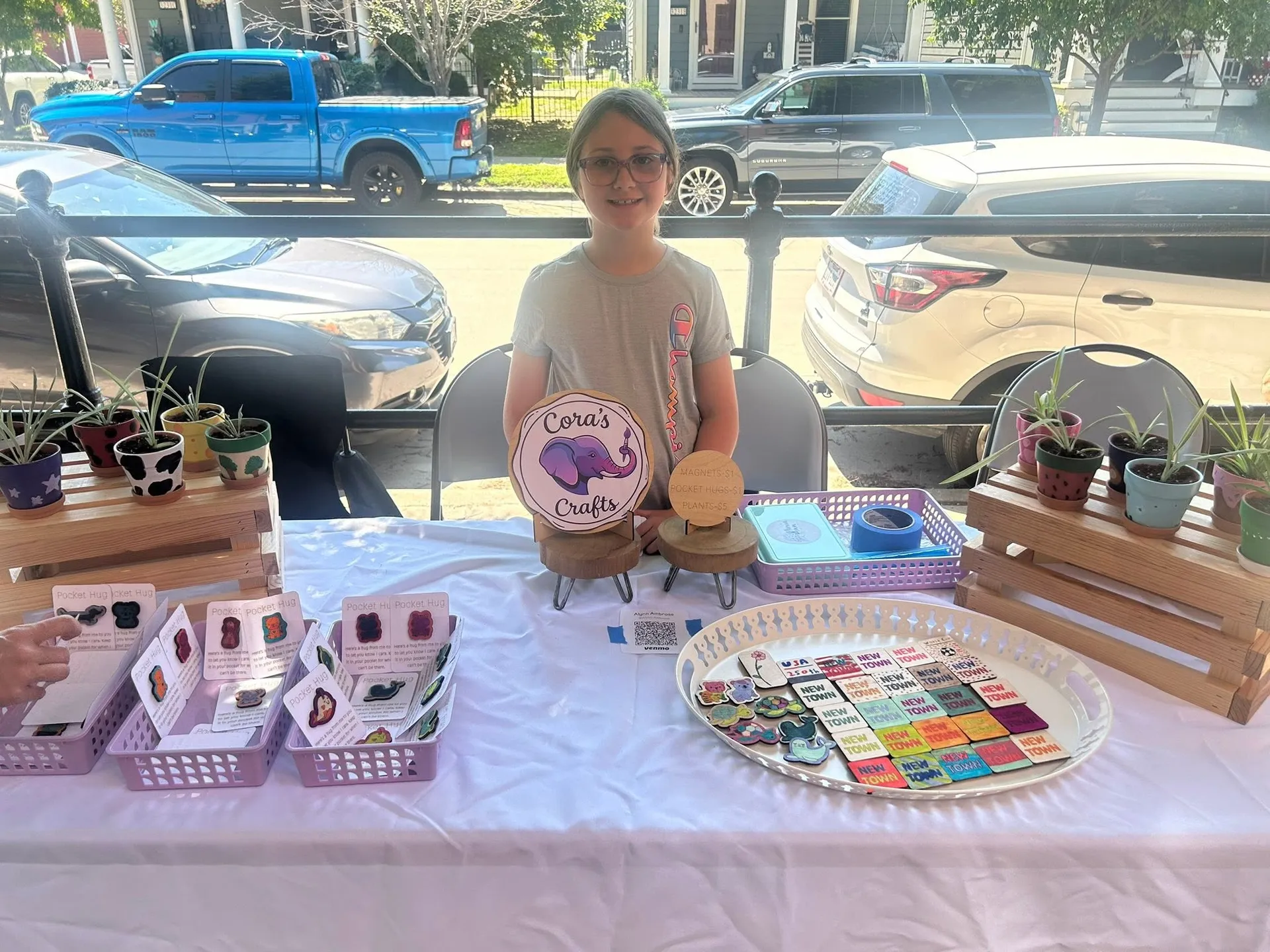 A young crafter displays handmade magnets, pocket hugs and painted plant pots at her Cora's Crafts table