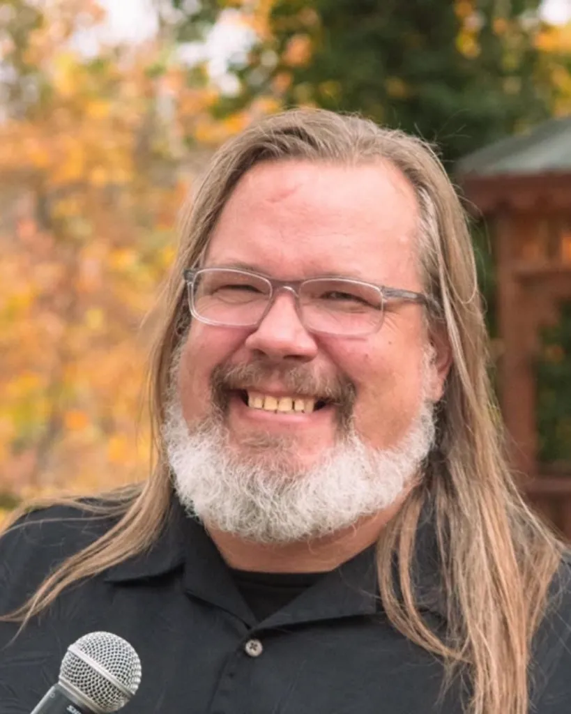 Tim Eveker smiling at a microphone with fall foliage behind him