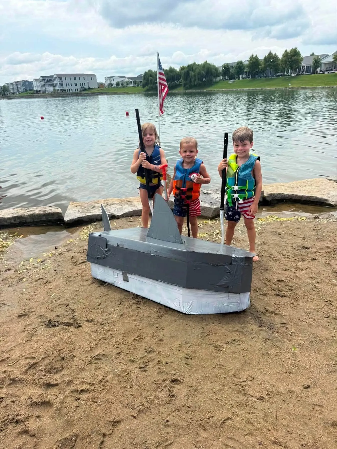 Three kids in life jackets pose with their shark-themed cardboard boat on the beach
