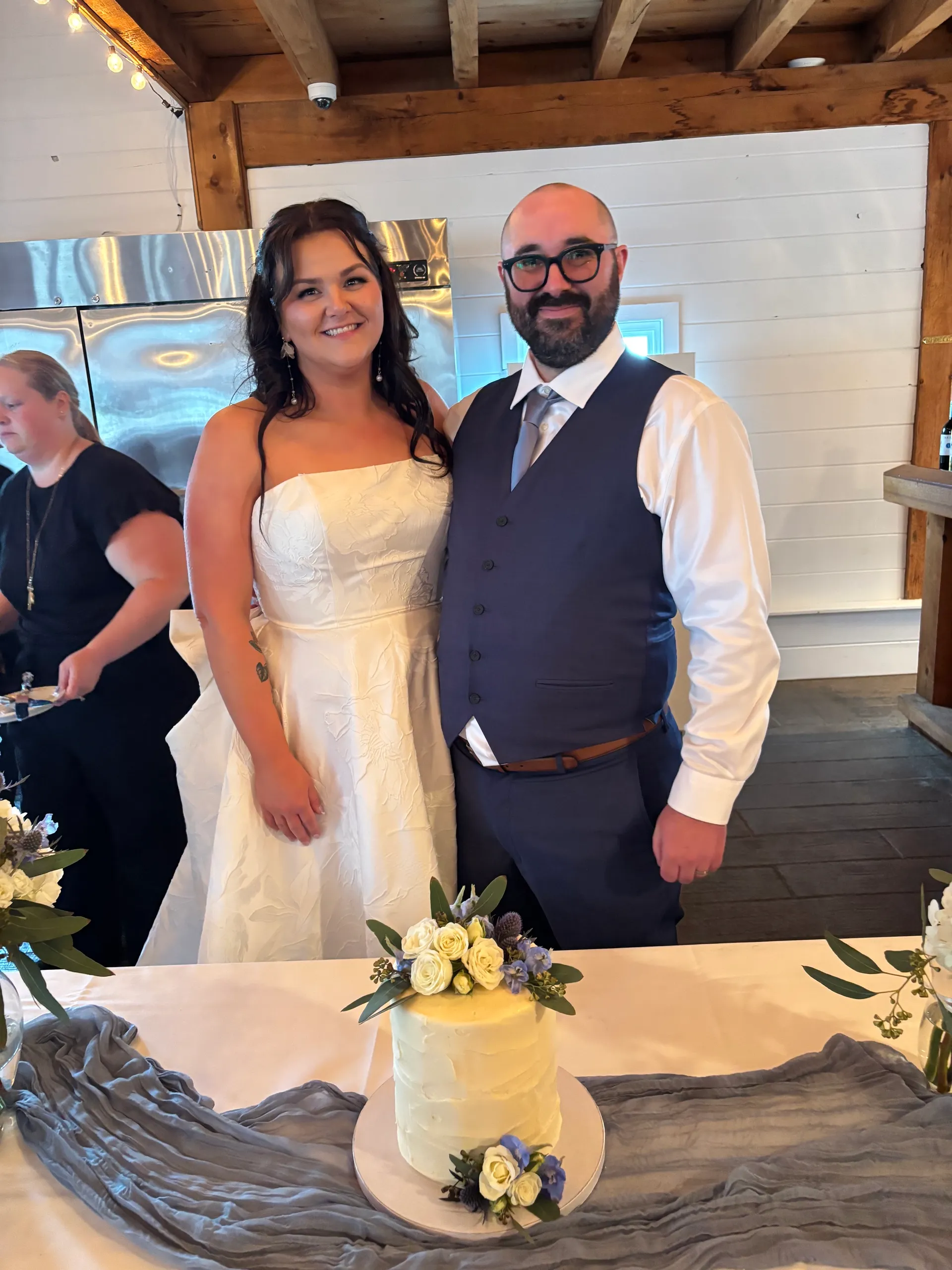 Jessica and John smiling behind their flower-topped wedding cake at the reception
