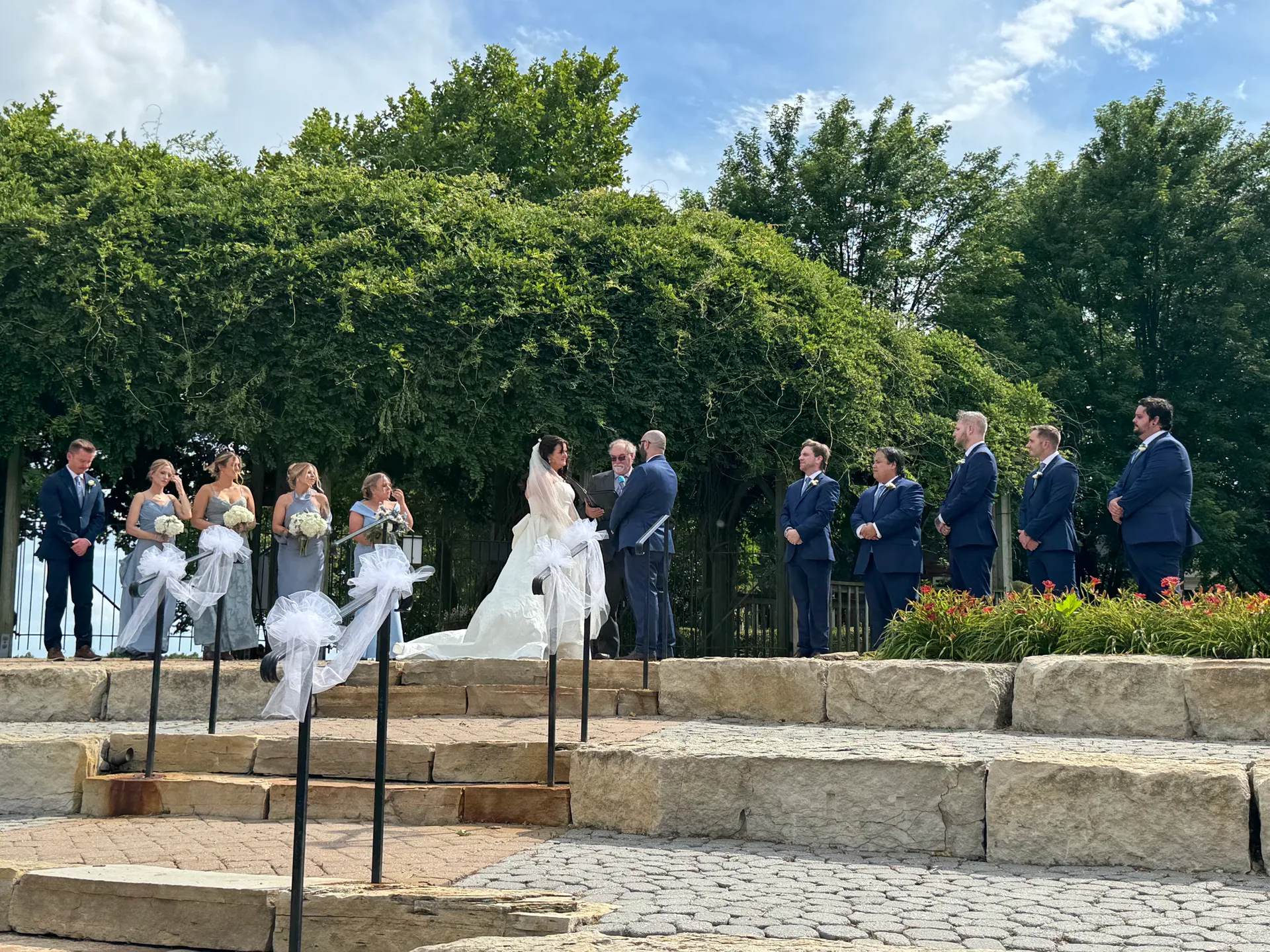 Jessica and John exchanging vows outdoors with their wedding party under lush greenery