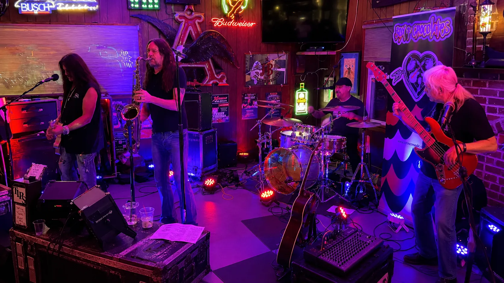 Box of Chocolates performing live on stage with vocals, saxophone, drums, and bass under colorful lights