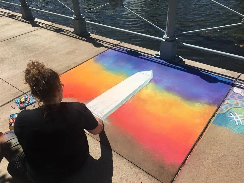 Lindsay Sutton chalking a colorful sunset design on the sidewalk beside the canal at the inaugural Chalk the Walk in 2018