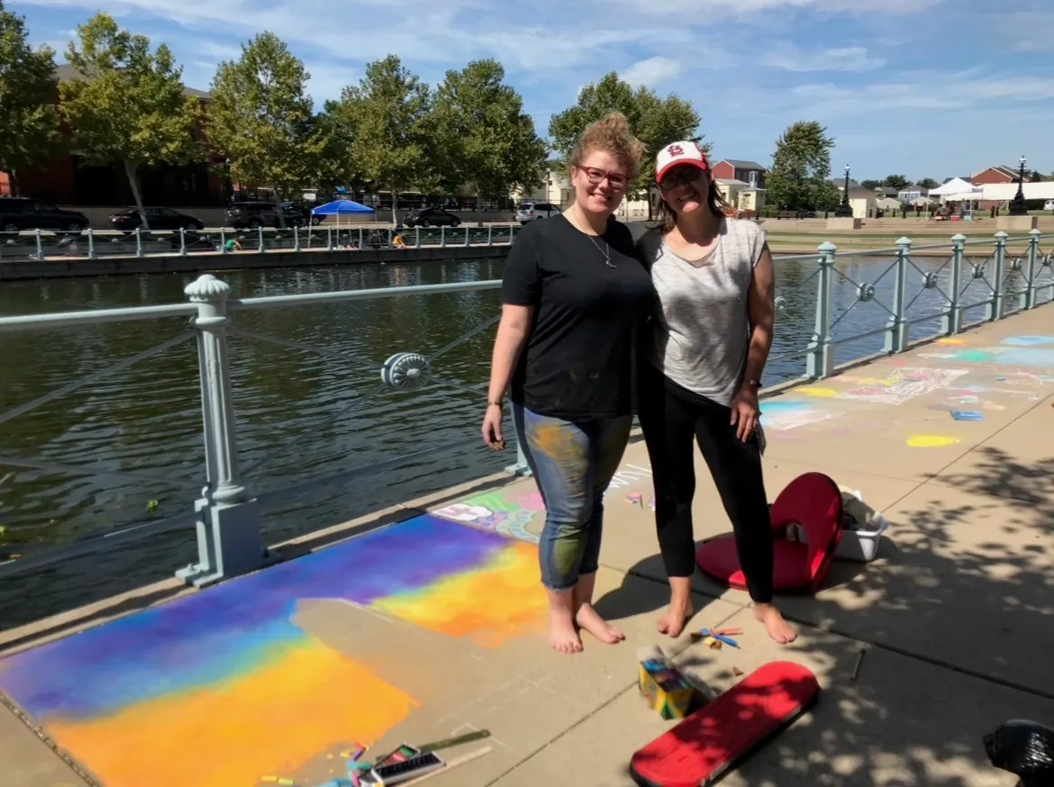 Two chalk artists smiling beside their sidewalk artwork along the canal at Chalk the Walk in New Town