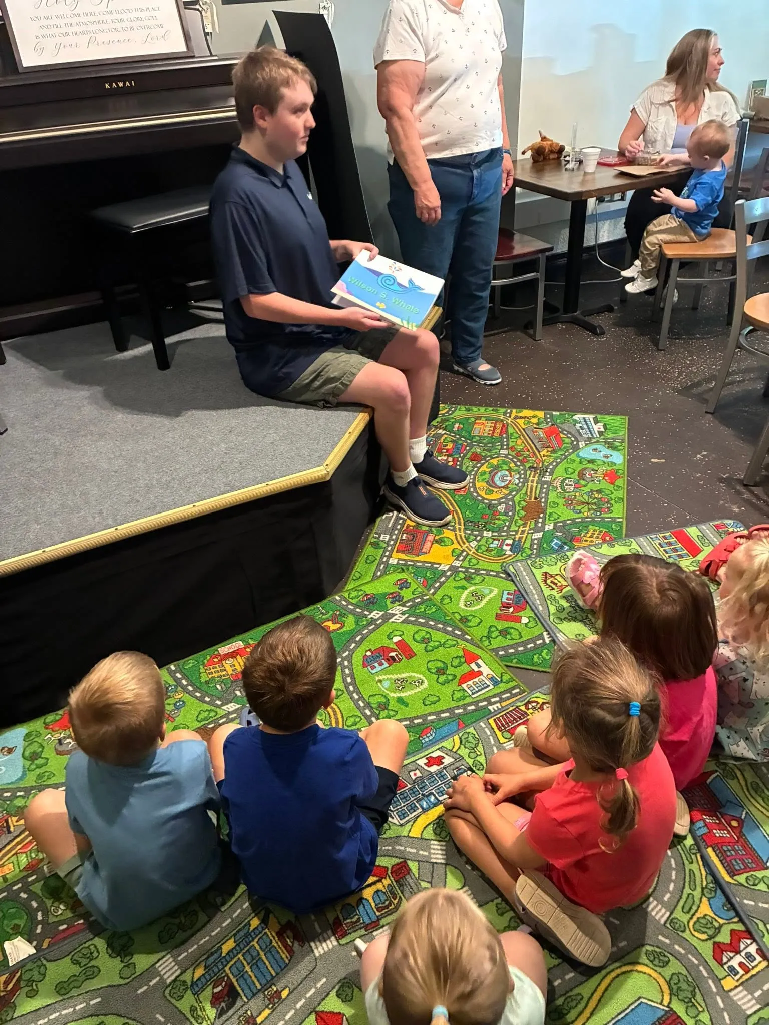 A young reader holds a picture book for children seated on colorful play rugs during Storytime at The Bridge Coffee House